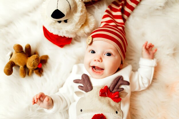 Małe dziecko w swetrze z jeleniem i czerwonym kapeluszem leży na miękkim białym kocu
