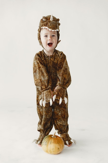 Mały chłopiec ubrany w brązowy kostium dinozaura trzymającego dynię. Chłopiec ma kaptur z twarzą dinozaura