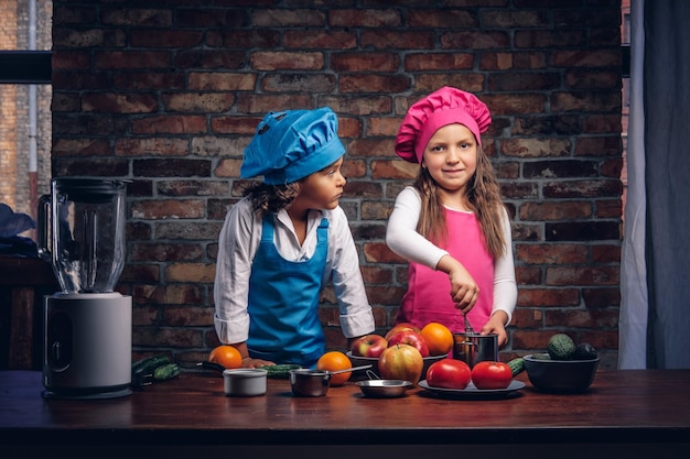 Mały chłopiec z brązowymi kręconymi włosami, ubrany w niebieski mundur kucharza i piękna dziewczyna ubrana w różowy mundur kucharza, wspólne gotowanie w kuchni przy ścianie z cegły. Cute little para kucharz.