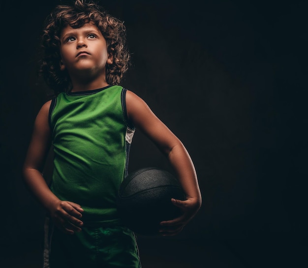 Mały koszykarz w sportowej trzymając piłkę w studio. Na białym tle na ciemnym tle z teksturą.