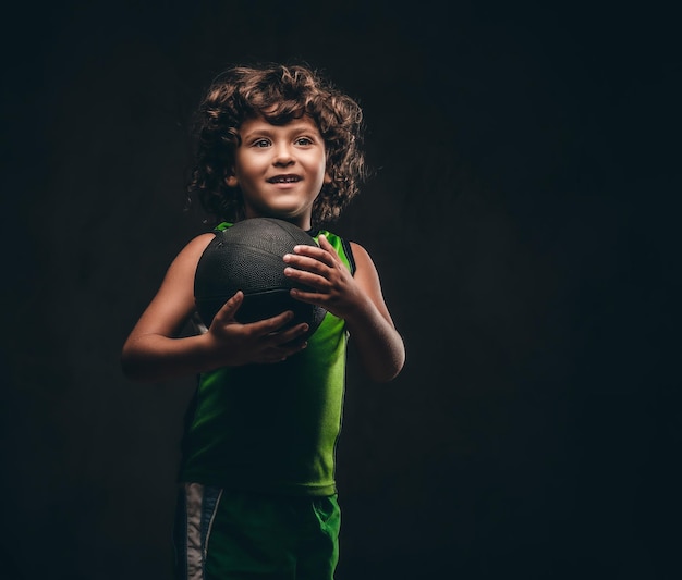 Mały Koszykarz W Sportowej Trzymając Piłkę W Studio. Na Białym Tle Na Ciemnym Tle Z Teksturą.