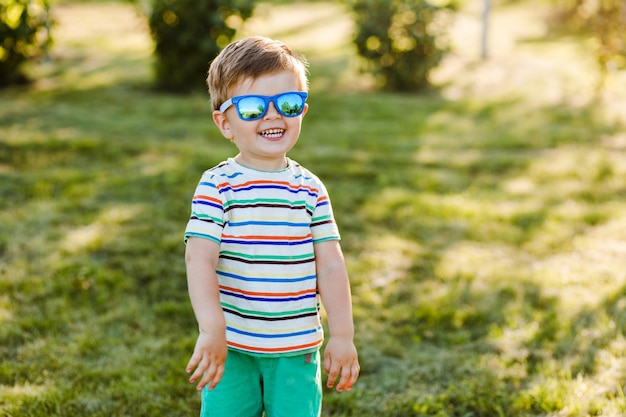 Mały ładny chłopiec uśmiecha się w letnim ogrodzie w jasnych okularach przeciwsłonecznych i pokazuje swoje szczęście.