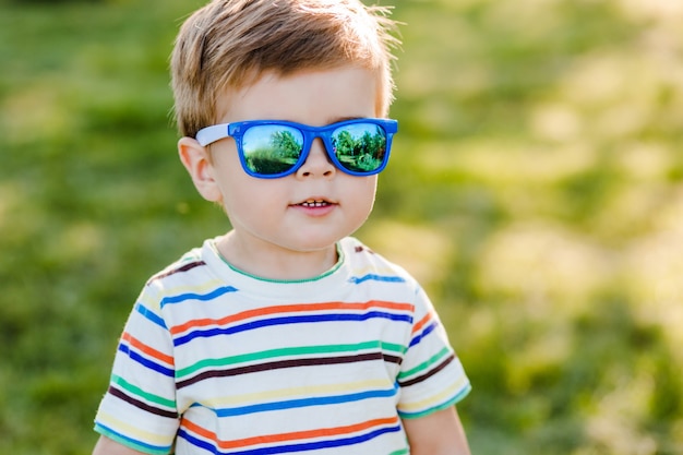 Mały śliczny chłopiec przebywa w ogrodzie w jasne okulary przeciwsłoneczne i uśmiech.