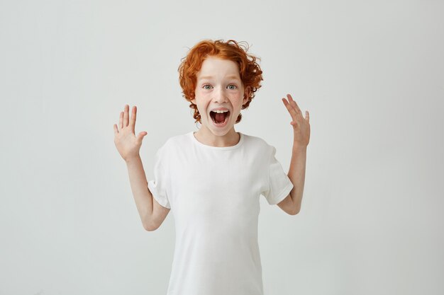 Mały śliczny rudy chłopiec z piegami w białej koszulce jest bardzo zaskoczony i szczęśliwy