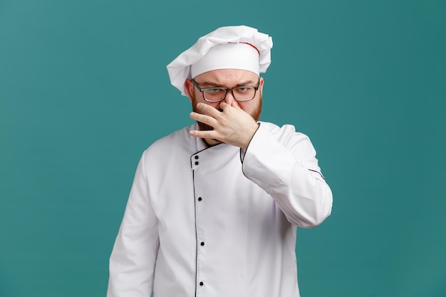 Marszcząc brwi, młody mężczyzna szef kuchni w mundurze okularowym i czapce, patrząc na kamerę pokazującą gest nieprzyjemnego zapachu na białym tle na niebieskim tle