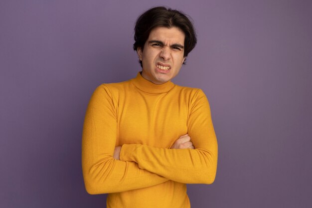 Marszcząc brwi, młody przystojny facet ubrany w żółty sweter z golfem, krzyżujący ręce izolowane na fioletowej ścianie