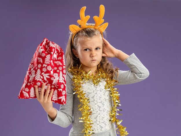 Marszcząca brwi dziewczynka ubrana w świąteczny obręcz do włosów z girlandą na szyi trzyma świąteczną torbę kładąc rękę na głowie na białym tle na niebieskiej ścianie