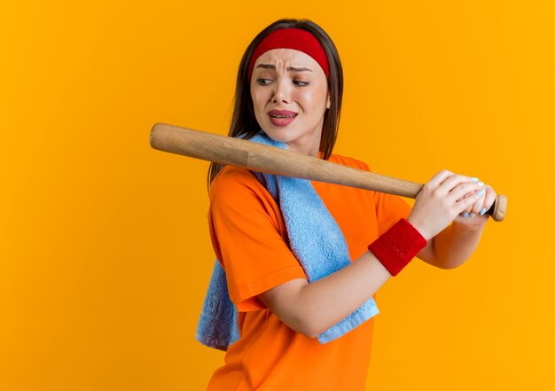 Marszcząca brwi, młoda sportowa kobieta nosząca opaskę i opaski na nadgarstkach z ręcznikiem na ramieniu trzymająca kij bejsbolowy patrząc z boku na białym tle na pomarańczowej ścianie z miejscem na kopię