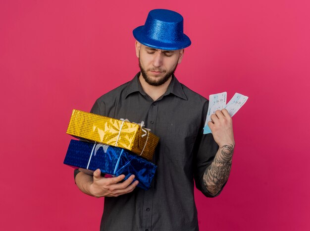 Marszczący brwi brwi młody przystojny słowiański facet w kapeluszu imprezowym, trzymając i patrząc na paczki z prezentami, trzymając bilety lotnicze na białym tle na szkarłatnym tle