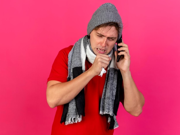 Marszczący brwi młody przystojny blondyn chory w czapce zimowej i szaliku rozmawia przez telefon trzymając serwetkę trzymając rękę w pobliżu ust, kaszel patrząc w dół odizolowany na różowej ścianie z miejscem na kopię