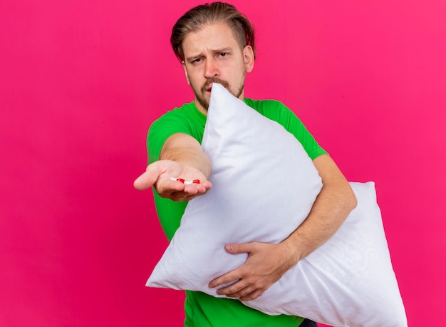 Bezpłatne zdjęcie marszczący brwi młody przystojny słowiański chory mężczyzna przytulający poduszkę wyciągający kapsułki do przodu patrząc na przód odizolowany na różowej ścianie z miejscem na kopię