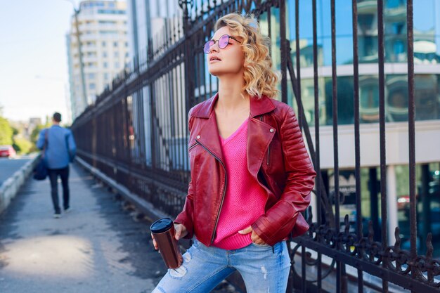 Marzycielska blond dziewczyna spacerująca po ulicach, pijąca kawę lub cappuccino. Stylowy jesienny strój, skórzana kurtka i dzianinowy sweter. Różowe okulary przeciwsłoneczne.