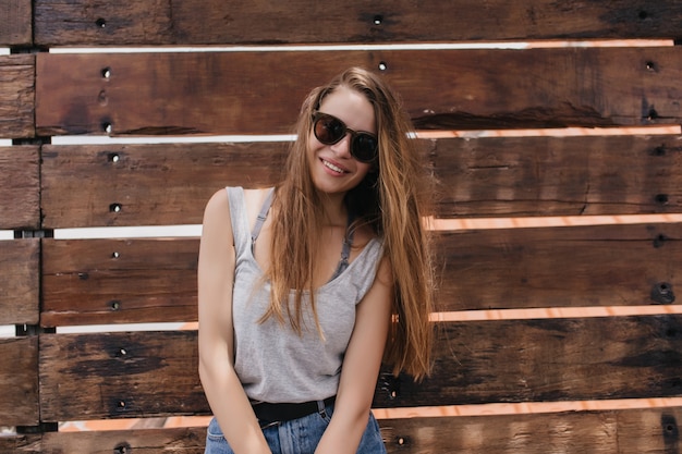 Bezpłatne zdjęcie marzycielska dziewczyna z długimi brązowymi włosami pozuje w pobliżu drewnianej ściany z uroczym uśmiechem. portret beztroski model europejski w letnich ubraniach.