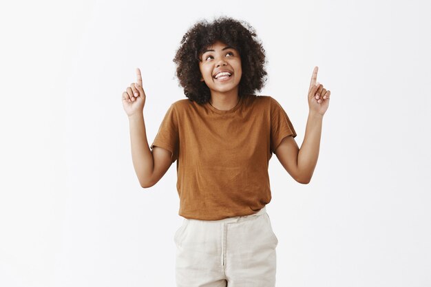 marzycielska i kreatywna atrakcyjna ciemnoskóra modelka z fryzurą afro w modnym stroju, spoglądająca i wskazująca w górę z radosnym rozbawionym uśmiechem, patrząc na interesującą przestrzeń do kopiowania