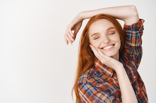 Marzycielska nastolatka z rudymi naturalnymi włosami, pozująca na białej ścianie, uśmiechnięta szczęśliwa z zamkniętymi oczami, dotykająca idealnej twarzy bez makijażu