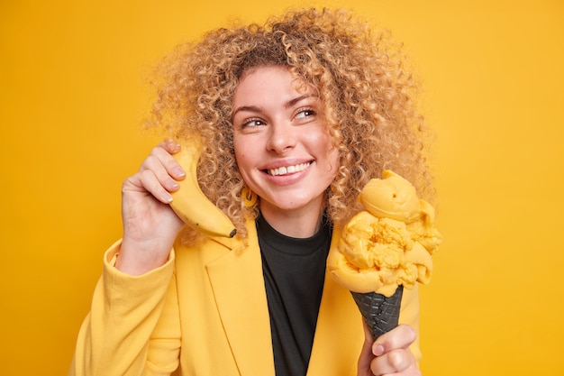 Marzycielska uśmiechnięta kobieta z kręconymi włosami odwraca wzrok w zamyśleniu trzyma lody w kształcie stożka zjada mrożony deser trzyma banana w pobliżu ucha udaje, że rozmowa telefoniczna jest w dobrym nastroju żółta ściana