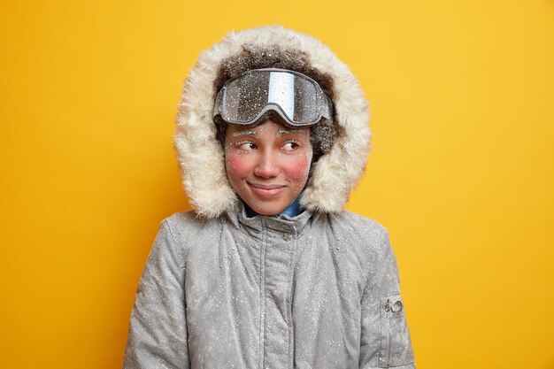 Marzycielska zimowa dziewczyna z czerwoną zmarzniętą twarzą cieszy się wakacjami w górskim kurorcie podczas mroźnego, śnieżnego dnia przykryta płatkami śniegu ubrana w ciepłą kurtkę z kapturem nosi gogle narciarskie lubi sporty ekstremalne.