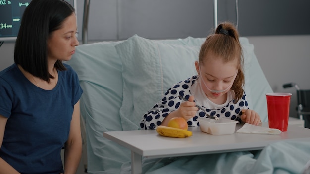 Bezpłatne zdjęcie matka siedzi obok chorej córki podczas jedzenia obiadu, zdrowienia po operacji. dziecko hospitalizowane o zdrowej żywności odżywianie śniadanie na oddziale szpitalnym podczas badania