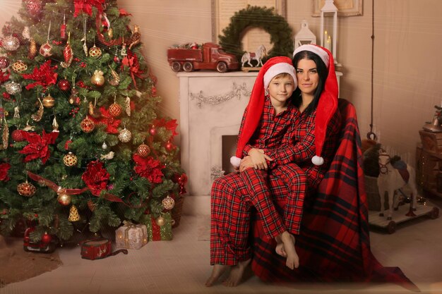 Matka syna w świątecznej piżamie przytulanie mikołaja siedzącego na krześle przy kominku