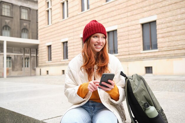 Bezpłatne zdjęcie media społecznościowe i ludzie, młoda ruda dziewczyna siedząca na ulicy, korzysta z aplikacji na telefon komórkowy, wyszukuje informacje