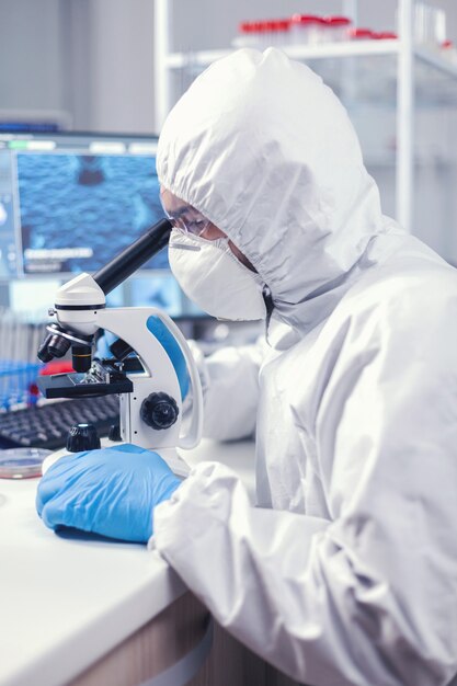 Medyk w czasie globalnej pandemii pracujący na mikroskopie ubrany w kombinezon ochronny