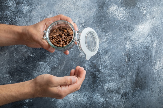 Męska ręka trzyma szklany słoik aromatycznych ziaren kawy na marmurowym tle