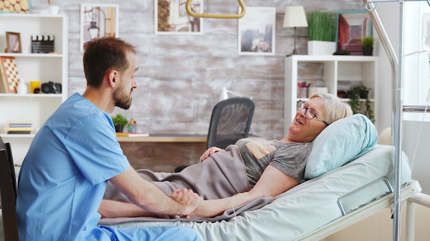 Męski opiekun rozmawiający z chorą starszą panią leżącą w szpitalnym łóżku, bierze kobietę za rękę