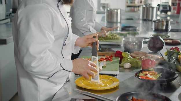 Bezpłatne zdjęcie męski szef kuchni tłuczący parmezan na tarce kuchennej, aby ugotować przepis gastronomiczny w profesjonalnej kuchni. autentyczny kucharz przygotowuje rozdrobniony składnik cheddar, aby przygotować danie dla smakoszy.