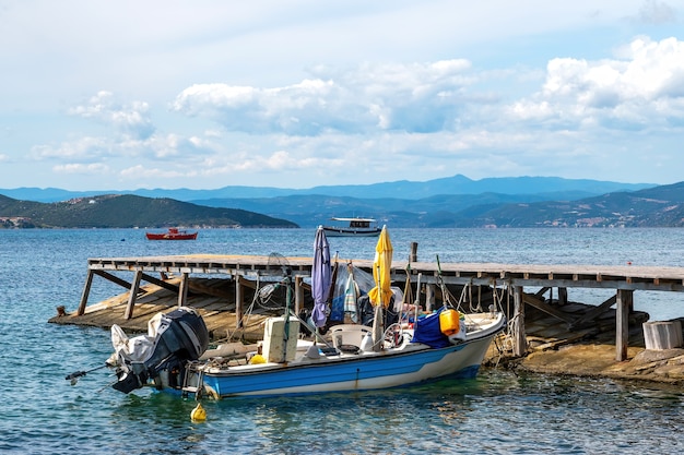 Metalowa, kolorowa łódź motorowa na plaży na molo na wybrzeżu Morza Egejskiego, wzgórzach i mieście w Ouranoupolis, Grecja