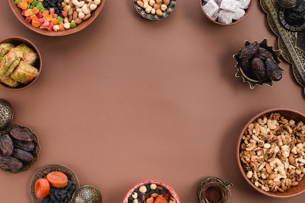 Metalowe i gliniane miseczki suszonych owoców; Daktyle; lukum; orzechy i baklava ułożone w okrągły kształt na brązowym tle