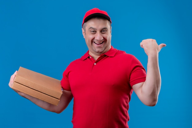 Mężczyzna dostawy ubrany w czerwony mundur i czapkę, trzymając pudełka po pizzy, uśmiechając się ze szczęśliwą twarzą, podnosząc pięść po zwycięstwie stojącym nad odizolowaną niebieską przestrzenią