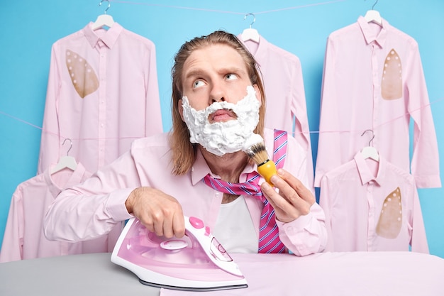 Bezpłatne zdjęcie mężczyzna goli i prasuje ubrania w tym samym czasie używa żelazka elektrycznego pozuje w pralni ubiera się na specjalne okazje. koncepcja pracy domowej