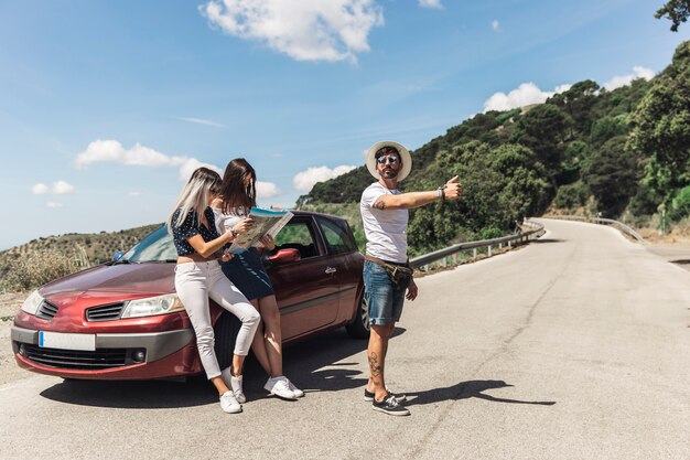 Mężczyzna hitchhiking na drogowej pozyci przed żeńskimi przyjaciółmi patrzeje mapę