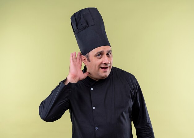 Mężczyzna kucharz na sobie czarny mundur i kapelusz kucharz, trzymając rękę przy uchu, próbując słuchać rozmowy kogoś stojącego na zielonym tle