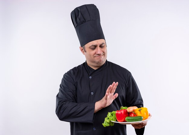 Mężczyzna kucharz na sobie czarny mundur i kapelusz kucharz trzymając talerz ze świeżymi warzywami, czyniąc gest obrony ze sceptycznym wyrazem twarzy stojącej na białym tle