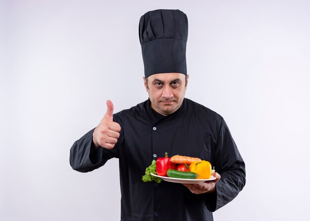 Mężczyzna kucharz na sobie czarny mundur i kapelusz kucharz trzymając talerz ze świeżymi warzywami pokazując kciuk do góry stojących na białym tle