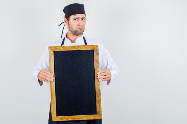 Mężczyzna kucharz trzyma tablicę w mundurze, fartuchu i wygląda na niezadowolonego. przedni widok.