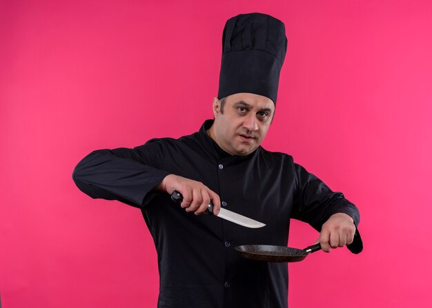Mężczyzna kucharz ubrany w czarny mundur i kapelusz kucharz trzyma patelnię i nóż patrząc na kamery z poważną twarzą stojącą na różowym tle