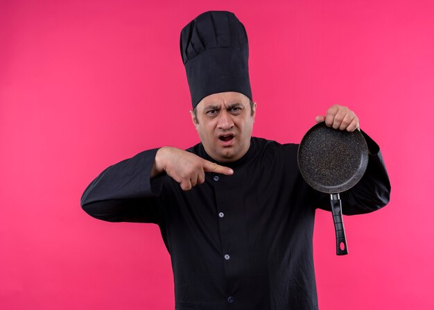 Bezpłatne zdjęcie mężczyzna kucharz ubrany w czarny mundur i kapelusz kucharza trzyma patelnię wskazując palcem na nią, krzycząc z agresywnym wyrazem stojącym na różowym tle