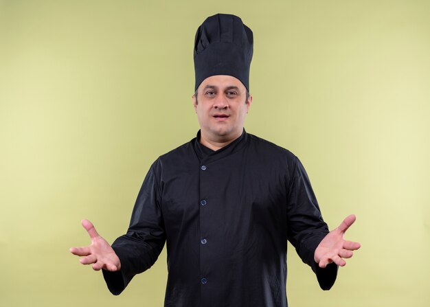 Mężczyzna kucharz ubrany w czarny mundur i kapelusz kucharza wygląda na niezadowolonego, trzymając ręce na zewnątrz, zadając pytanie stojąc na zielonym tle