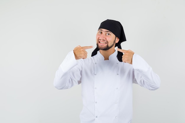 Bezpłatne zdjęcie mężczyzna kucharz wskazując na jego uśmiech w białym mundurze i patrząc zadowolony, widok z przodu.