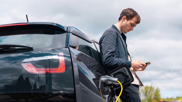 Mężczyzna ładujący swój samochód elektryczny na stacji ładującej i używający smartfona