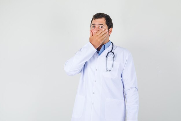 Mężczyzna lekarz obejmujący usta ręką w białym fartuchu i wyglądający na zaskoczonego. przedni widok.