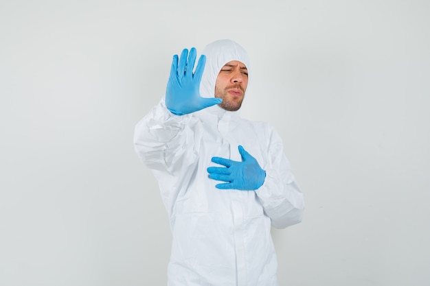 Mężczyzna lekarz pokazując gest stop i trzymając rękę na klatce piersiowej w kombinezonie ochronnym