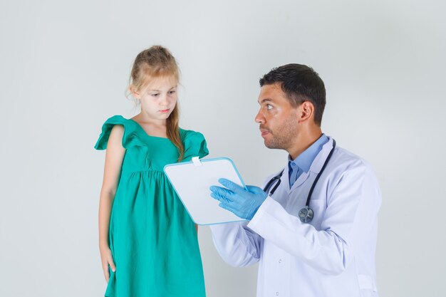 Mężczyzna lekarz pokazując małą dziewczynkę coś na pokładzie w białym fartuchu widok z przodu.