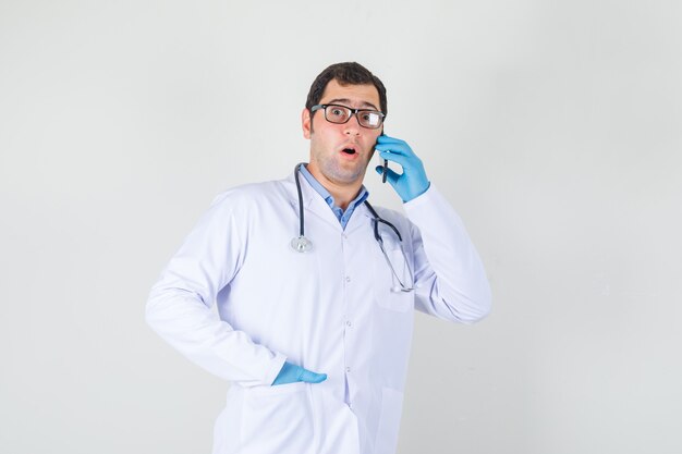 Mężczyzna lekarz rozmawia przez telefon ręką w kieszeni w białym fartuchu, rękawiczkach, okularach i wygląda na zaskoczonego