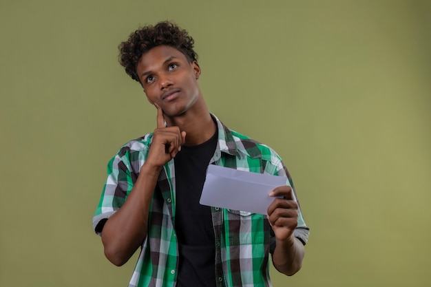 Mężczyzna Młody Podróżnik African American Gospodarstwa List Patrząc Z Palcem Na Brodzie Z Zamyślonym Wyrazem Myśli Stojącej Na Zielonym Tle