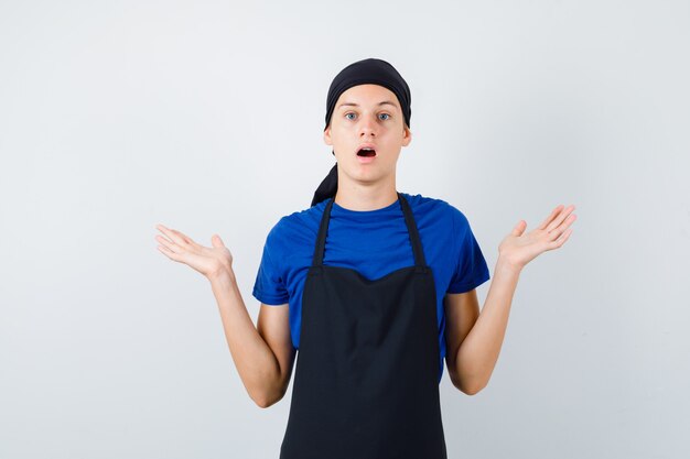 Mężczyzna nastolatek kucharz wzruszając ramionami w koszulce, fartuchu i patrząc w szoku. przedni widok.