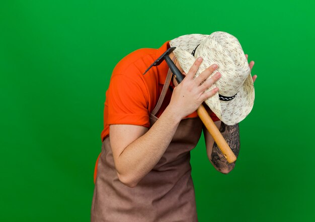 Mężczyzna ogrodnik w kapeluszu ogrodniczym kładzie ręce na głowie, trzymając grabie