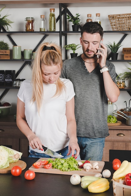 Mężczyzna opowiada na telefonie komórkowym patrzeje żony tnących warzywa z nożem w kuchni